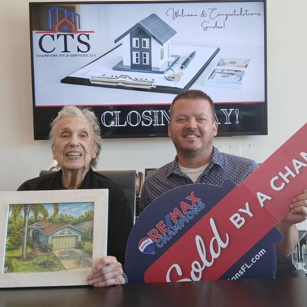 Keith Mathias, a RE/MAX Champions realtor, smiles with an elderly client in New Port Richey after successfully selling her home. They are seated in front of a "Closing Day" sign at Champions Title Services.
